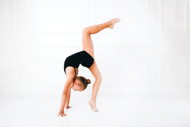 jong meisje in een zwart jurk doen gymnastiek staande op een been op witte achtergrond - Foto, afbeelding
