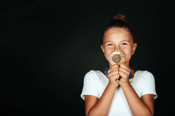Portrait of little gymnast girl in white bodysuit happy showing, holding her medal isolated on black background - Фото, изображение