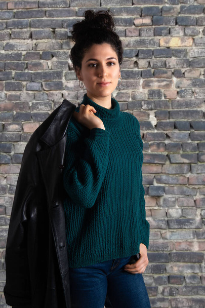 Portrait of young attractive woman in sweater with leather jacket on her shoulder. Brunette female model looking at camera. Old brick wall background - Fotoğraf, Görsel