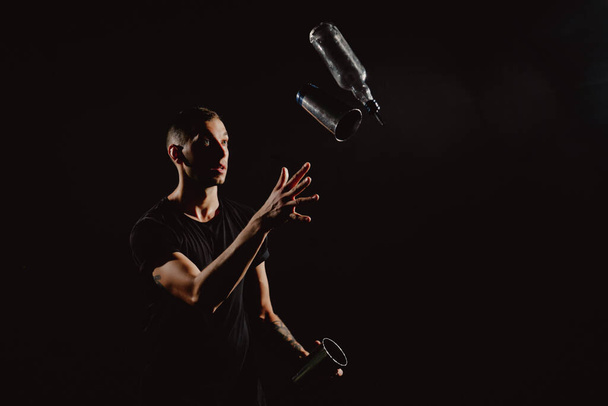 Δραματικό πορτραίτο ενός όμορφου μπάρμαν που κουνάει και ανακατεύει κοκτέιλ αλκοόλ. Επαγγελματίας μπάρμαν ταχυδακτυλουργός και μπουκάλι σε χέρια που απομονώνονται σε μαύρο - Φωτογραφία, εικόνα