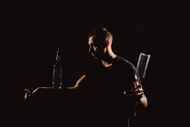 Dramatyczny portret przystojnego barmana trzęsącego się i mieszającego koktajl alkoholowy. Profesjonalny barman żonglerka shaker i butelka w rękach izolowane na czarno - Zdjęcie, obraz