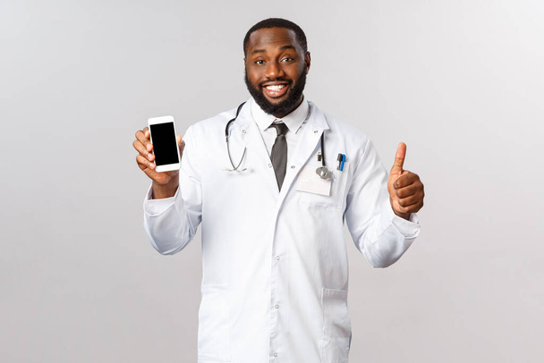 Covid19, pandemian ja verkkotapaamisen käsite. Komea hymyilevä afrikkalais-amerikkalainen lääkäri suosittelee käyttämään mobiilisovellusta potilaiden hoitoon, soittamaan lääkärille ja neuvottelemaan hoidosta tai neuvottelemaan
 - Valokuva, kuva