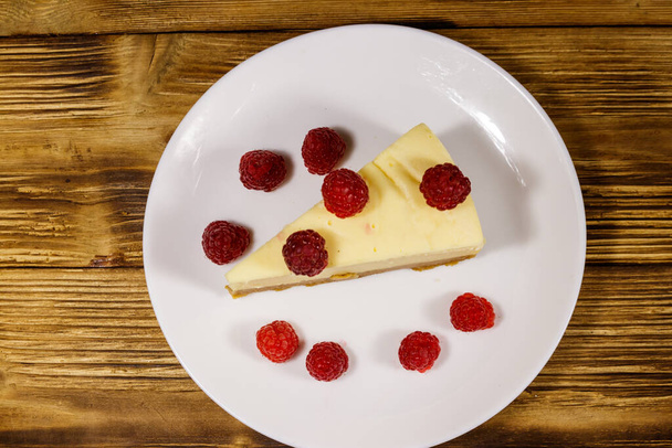Un pedazo de sabroso pastel de queso de Nueva York con frambuesas en un plato blanco sobre una mesa de madera. Vista superior - Foto, imagen