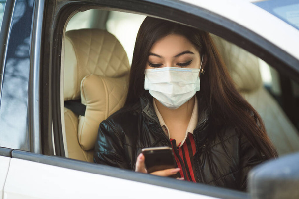 beautiful young woman in medical face mask in a car with a phone in her hand - Photo, image