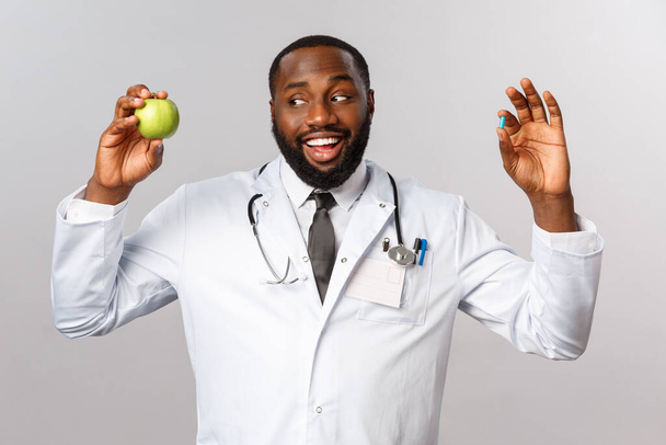 医療、医療、健康的なライフスタイルの概念。白いコートでフレンドリーなアフリカ系アメリカ人医師,リンゴと錠剤を保持,患者は薬なしで健康を維持示唆しています,より多くの果物を食べる - 写真・画像