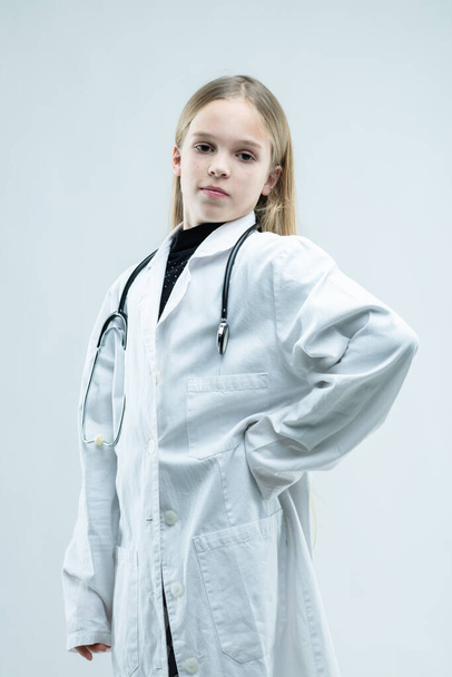 Proud young girl in a white lab coat playing at being a doctor or nurse with stethoscope around her neck isolated on white - 写真・画像