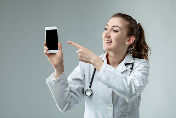 Жінка-лікар або медсестра в лабораторному пальто і стетоскоп вказує на порожній мобільний телефон з приємною посмішкою крупним планом на сірому фоні
 - Фото, зображення