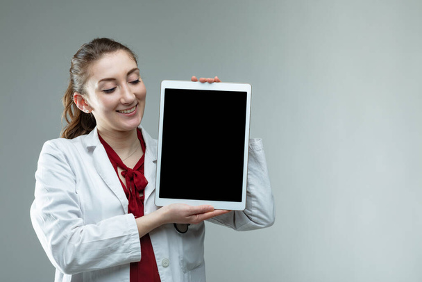 Улыбающаяся дружелюбная женщина врач или медсестра, держащая планшет на виду у зрителя на сером фоне с копировальным пространством
 - Фото, изображение