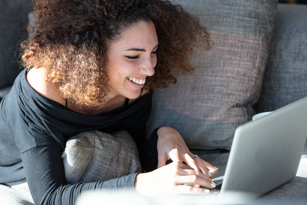 Mosolygó gyönyörű fiatal nő göndör barna hajjal, kényelmesen fekszik egy kanapén a laptopja előtt. Oldalsó portré fénymásoló felülettel - Fotó, kép