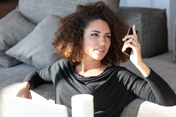 Młoda kobieta z trudem czyta na ekranie swojego telefonu, gdy relaksuje się na kanapie w domu z laptopem na słońcu, wykonując zdalną pracę dla swojej firmy. - Zdjęcie, obraz