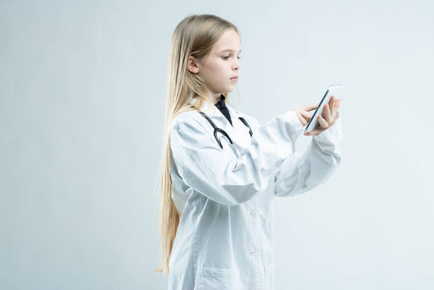 Молода дівчина, одягнена в медичну лабораторію пальто зі стетоскопом, прикидається лікарем, що стоїть, використовуючи свій мобільний телефон ізольовано на білому
 - Фото, зображення