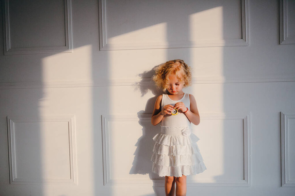 Uma menina tímida loira encaracolada em vestido branco brinca com pequeno relógio amarelo
 - Foto, Imagem