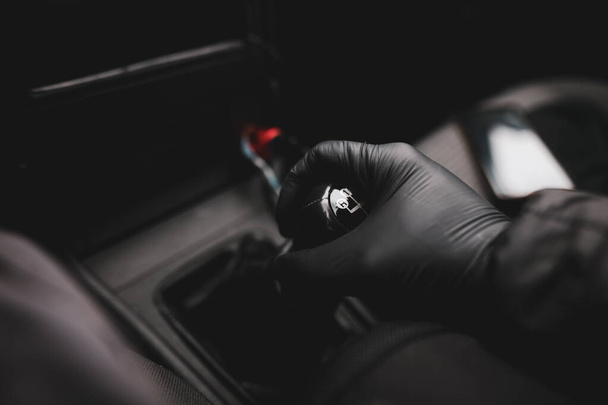 Λεπτομέρειες με το χέρι ενός οδηγού, σε ένα λαστιχένιο γάντι, σε ένα παλιό και σπασμένο πόμολο αλλαγής ταχυτήτων ενός χειροκίνητου κιβωτίου ταχυτήτων κατά τη διάρκεια της πανδημίας Covid-19. - Φωτογραφία, εικόνα