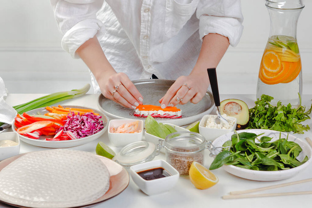 Woman's hands preparing the spring roll with fresh vegetables - putting carrots, cheese, basil and cucumber on rice paper. Appetizing diet asian dish. - Photo, Image