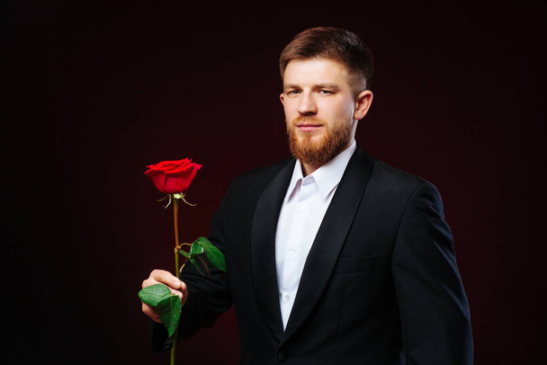 Hezký mladý muž v obleku stojí s červenou růží na černém pozadí. Romantická fotka muže držícího růži. - Fotografie, Obrázek