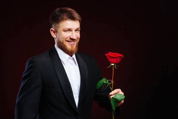 Bonito jovem de terno dando uma rosa vermelha em um fundo preto. Foto romântica do homem convidando uma mulher para sair
. - Foto, Imagem