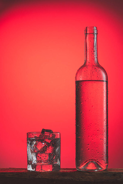 Orzeźwiający zimny napój w butelce z kroplami i szklanka z lodem na czerwonym tle. Koncepcja napojów orzeźwiających mrożonych. - Zdjęcie, obraz
