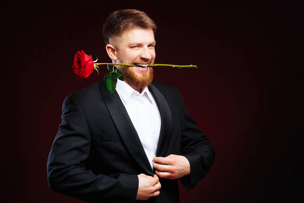 赤いバラを口に持つスーツ姿のロマンチックな青年が上着のボタンを押していた。情熱的な恋人. - 写真・画像