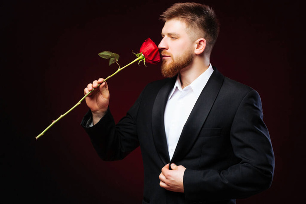 Romantic elegant man in suit is smeling the red rose on grey background. Getting ready for 8th of March. - Photo, Image