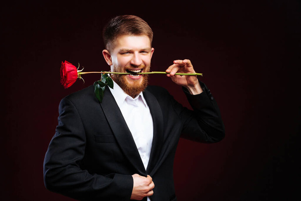 Романтичний молодий чоловік у костюмі тримає червону троянду в роті, застібаючи куртку. Пристрасний коханець
. - Фото, зображення