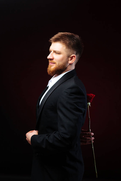 Homem bonito barbudo num fato que esconde uma rosa vermelha nas costas. Homem está preparando uma surpresa para sua namorada
. - Foto, Imagem