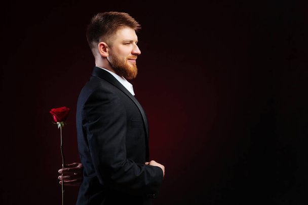 Ένας γενειοφόρος όμορφος άντρας με κοστούμι που κρύβει ένα κόκκινο τριαντάφυλλο πίσω από την πλάτη του. Ο άντρας ετοιμάζει μια έκπληξη για την κοπέλα του.. - Φωτογραφία, εικόνα