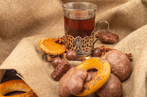 A faceted glass of tea in a vintage Cup holder, bagels, cookies, chocolate gingerbread and star anise on a background of rough-textured fabric. Close up - Photo, Image
