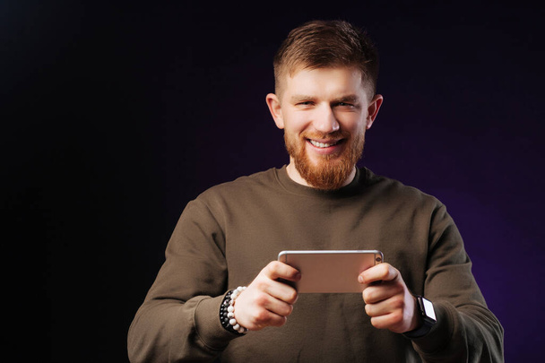 Χαμογελώντας γενειοφόρος νεαρός άνδρας σε πράσινο φούτερ παίζοντας παιχνίδια στο smartphone σε μωβ φόντο. - Φωτογραφία, εικόνα