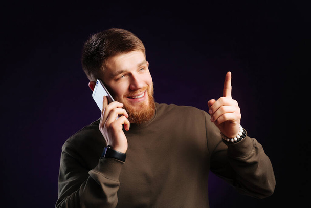 Homem barbudo jovem sorrindo e falando no telefone inteligente no estúdio no fundo escuro
. - Foto, Imagem