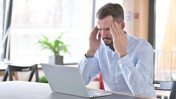 Hombre joven con portátil con dolor de cabeza en la oficina - Foto, Imagen