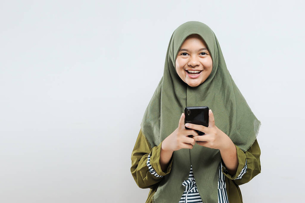 Mulher muçulmana bonita olhando para a câmera segurando telefone celular no fundo cinza
 - Foto, Imagem