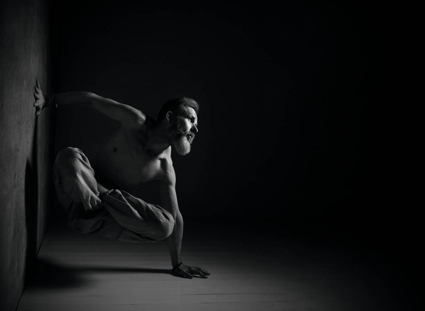 Aged man, naked torso, in brown pants is practicing yoga. Legs in lotus pose, setting one hand against wall, another one on floor - Photo, image