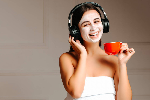 Mujer joven con mascarilla usando auriculares, escuchando música y tomando café. Joven mujer sonriente disfrutando de su mañana
. - Foto, Imagen
