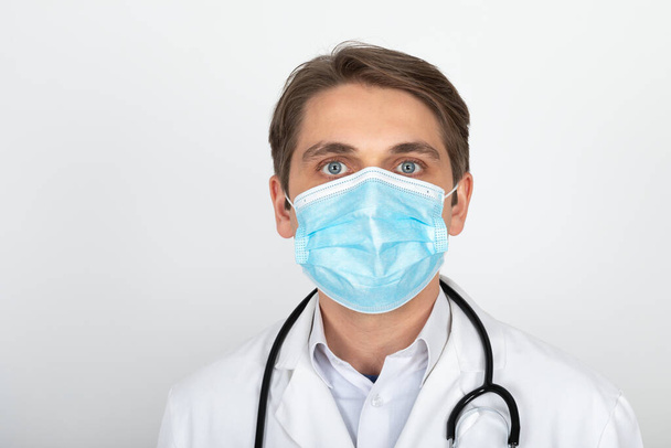 Handsome young physician wearing surgical mask is looking to the camera in front of white background - 写真・画像