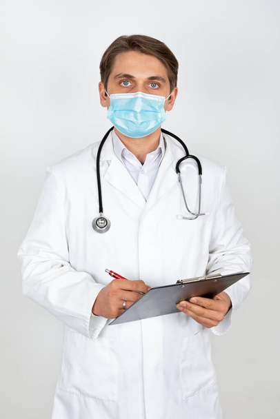 Handsome young physician wearing surgical mask is looking to the camera in front of white background - Valokuva, kuva