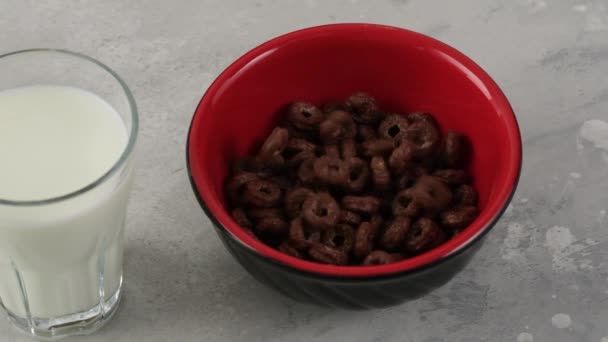Anillos de chocolate copos de maíz cayendo en tazón. Cereales de desayuno sobre mesa con vaso de leche
 - Metraje, vídeo