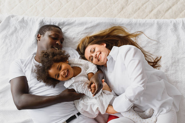 Portrait of happy multiracial young family lying on cozy white bed at home, smiling international mom and dad relaxing with little biracial girl child posing for picture in bedroom - Photo, image