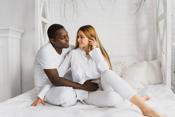 african american man hugging his charming girlfriend, sitting on white bed - Foto, afbeelding