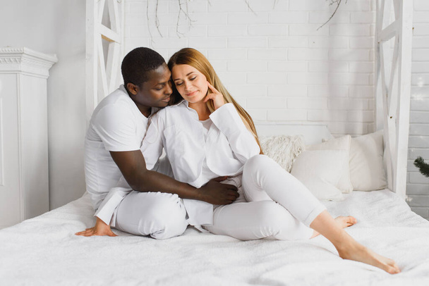 Portrait of happy multiracial young family lying on cozy white bed at home, smiling international mom and dad relaxing with little biracial girl child posing for picture in bedroom - Valokuva, kuva