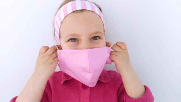 頭皮の小さな女の子は、コロナウイルスCOVID-19パンデミック白の背景に対して保護ピンクのファブリックマスクを身に着けています。子供の触れる顔、手作りの縫製保護マスク. - 映像、動画