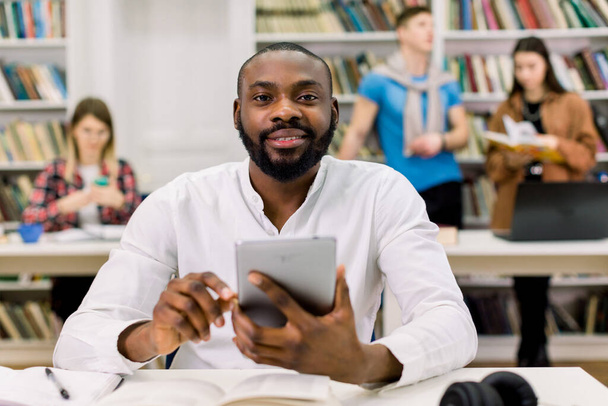 Happy young university multiracial students studying with books in library. Handsome African guy looking at camera while sitting at the table and working on tablet pc, i-pad - Foto, afbeelding