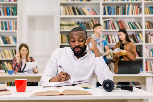 Young busy African man in white shirt, student studying in library at the university, talking on the smartphone and making notes in his copybook. Diverse students studying together on the background - Photo, image
