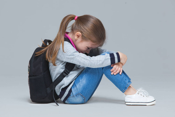 Primary school girl in jeans and uniform jacket is wearing backpack sitting tired after classes. Isolated on grey background - Foto, Imagen