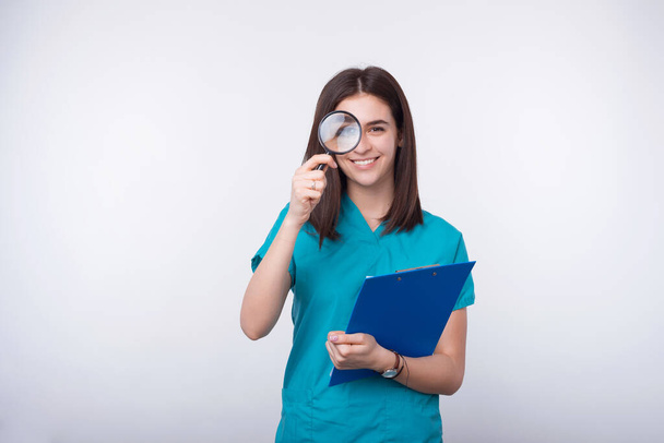 Cheerful young woman is looking through magnifying glass and holding a paper board on white background. - Foto, afbeelding
