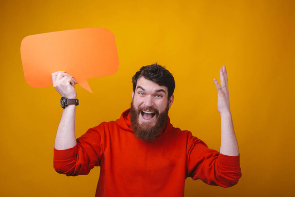 Bearded man is holding a speech bubble making blow your mind gesture on yellow background. - Фото, изображение