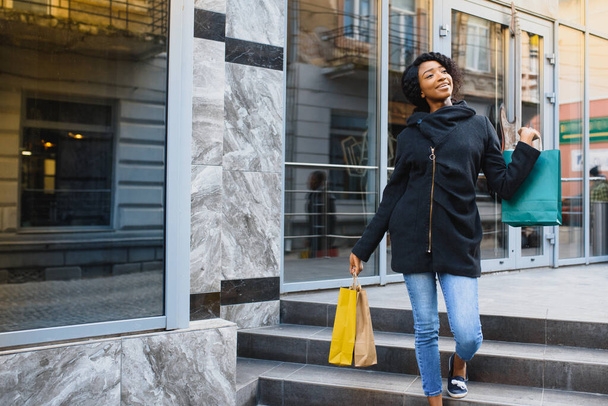Güzel kıvırcık saçlı Afro-Amerikan kadın elinde renkli alışveriş torbalarıyla bir alışveriş merkezinde duruyor.. - Fotoğraf, Görsel