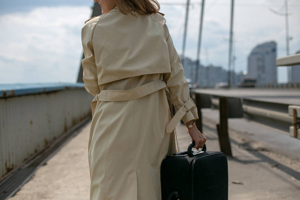 van streek jonge vrouw in een beige regenjas zwerft vermoeid met een koffer over de brug - Foto, afbeelding
