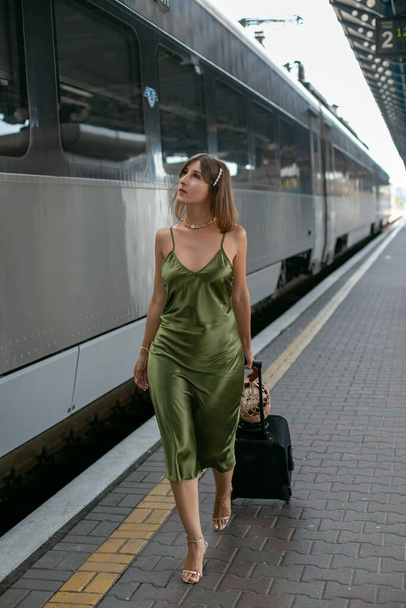 młoda brunetka w zielonej jedwabnej sukience spaceruje z walizką wzdłuż peronu pociągu - Zdjęcie, obraz