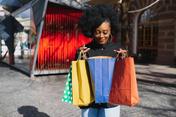 Attractive young African-American woman shopping - shopping bags, outdoors, street view, suitable for holiday shopping themes, among others - Photo, Image
