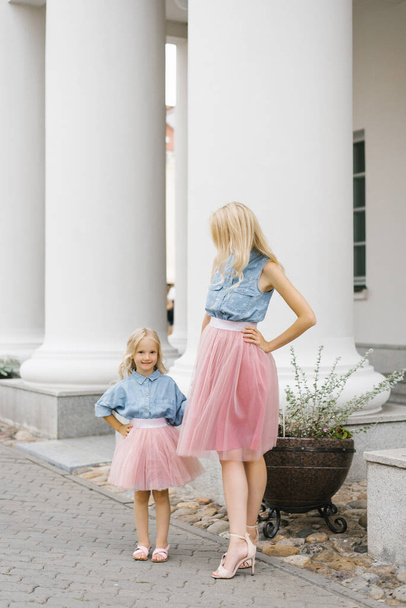 A beautiful young blonde mother and her charming daughter in the same romantic clothes stand near the columns and smile - Photo, Image